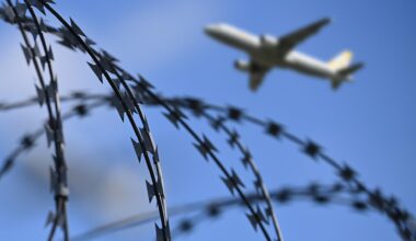 Ein startendes Passagierflugzeug ist am Himmel zu sehen, im Vordergrund ein Zaun mit Stacheldraht.