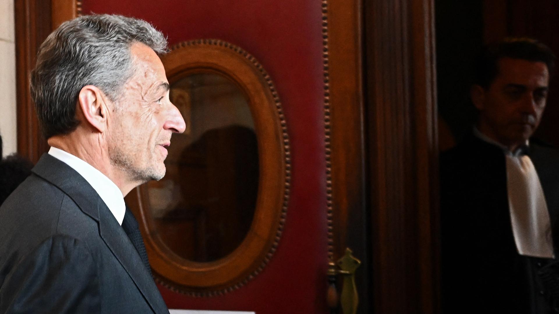 Sarkozy im schwarzen Jackett im Seitenprofil vor einer Wand mit zwei Spiegeln. Sarkozy im schwarzen Jackett im Seitenprofil vor einer Wand mit zwei Spiegeln.
