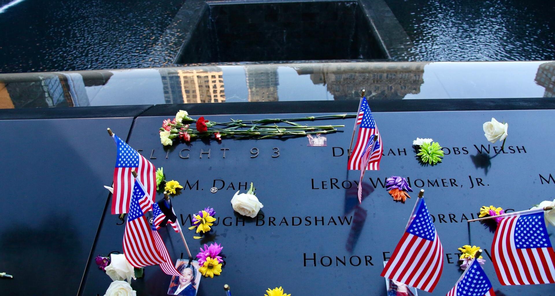 Das Bild zeigt eine Gedenktafel mit Namen der zahlreichen Opfer. Es liegen außerdem Blumen und US-amerikanische Flaggen darauf.