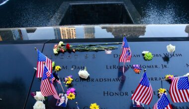 Das Bild zeigt eine Gedenktafel mit Namen der zahlreichen Opfer. Es liegen außerdem Blumen und US-amerikanische Flaggen darauf.