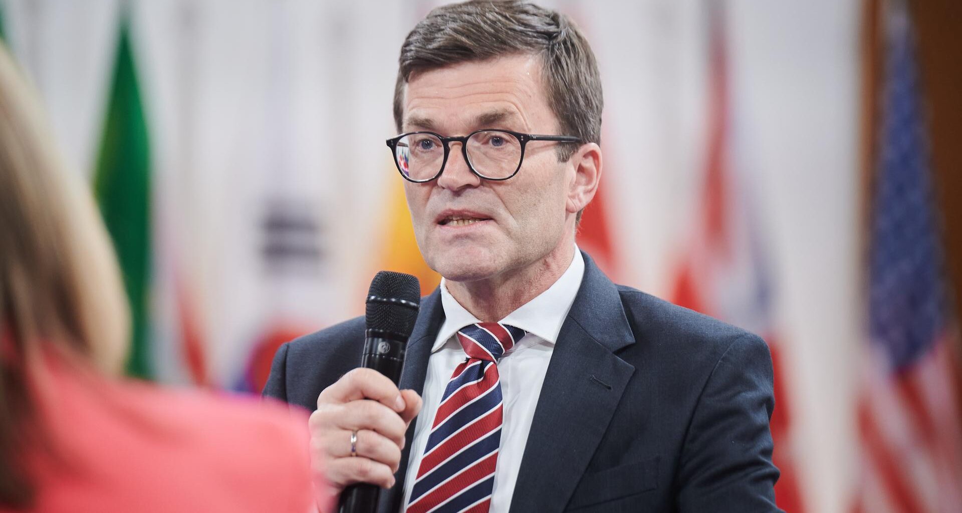 Thoms mit Brille, schwarzem Jackett und Krawatte hält ein Mikrofon und sagt etwas. Dahinter unscharf verschiedene Flaggen.