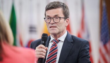 Thoms mit Brille, schwarzem Jackett und Krawatte hält ein Mikrofon und sagt etwas. Dahinter unscharf verschiedene Flaggen.