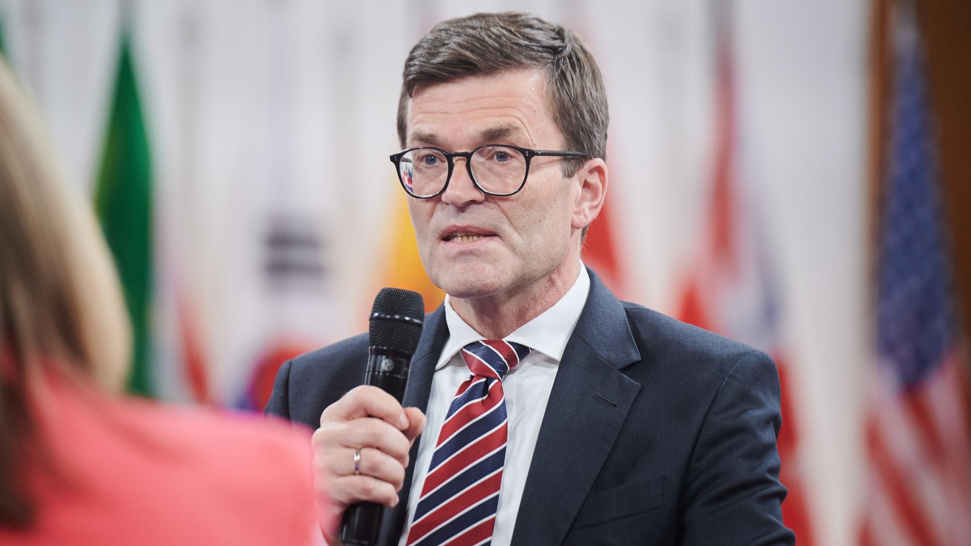 Thoms mit Brille, schwarzem Jackett und Krawatte hält ein Mikrofon und sagt etwas. Dahinter unscharf verschiedene Flaggen. 