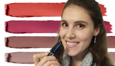 Eine Frau mit langen dunklen Haaren hält sich einen Lippenstift vors Gesicht. Im Hintergrund Streifen von Lippenstift in unterschiedlichen Farben