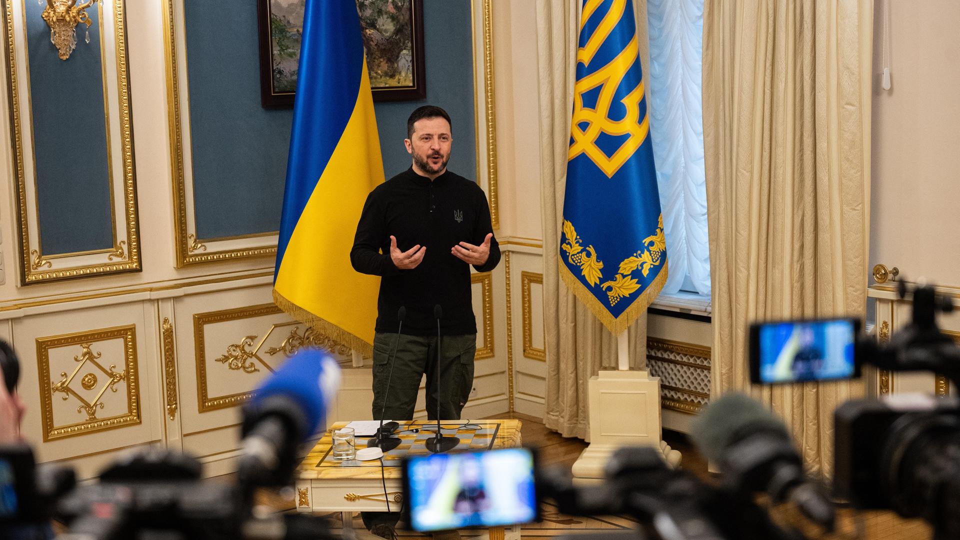 Der ukrainische Präsident Selenskyj steht vor mehreren Pressevertretern und gibt eine Erklärung ab, im Hintergrund sind zwei ukrainische Flaggen zu sehen. 