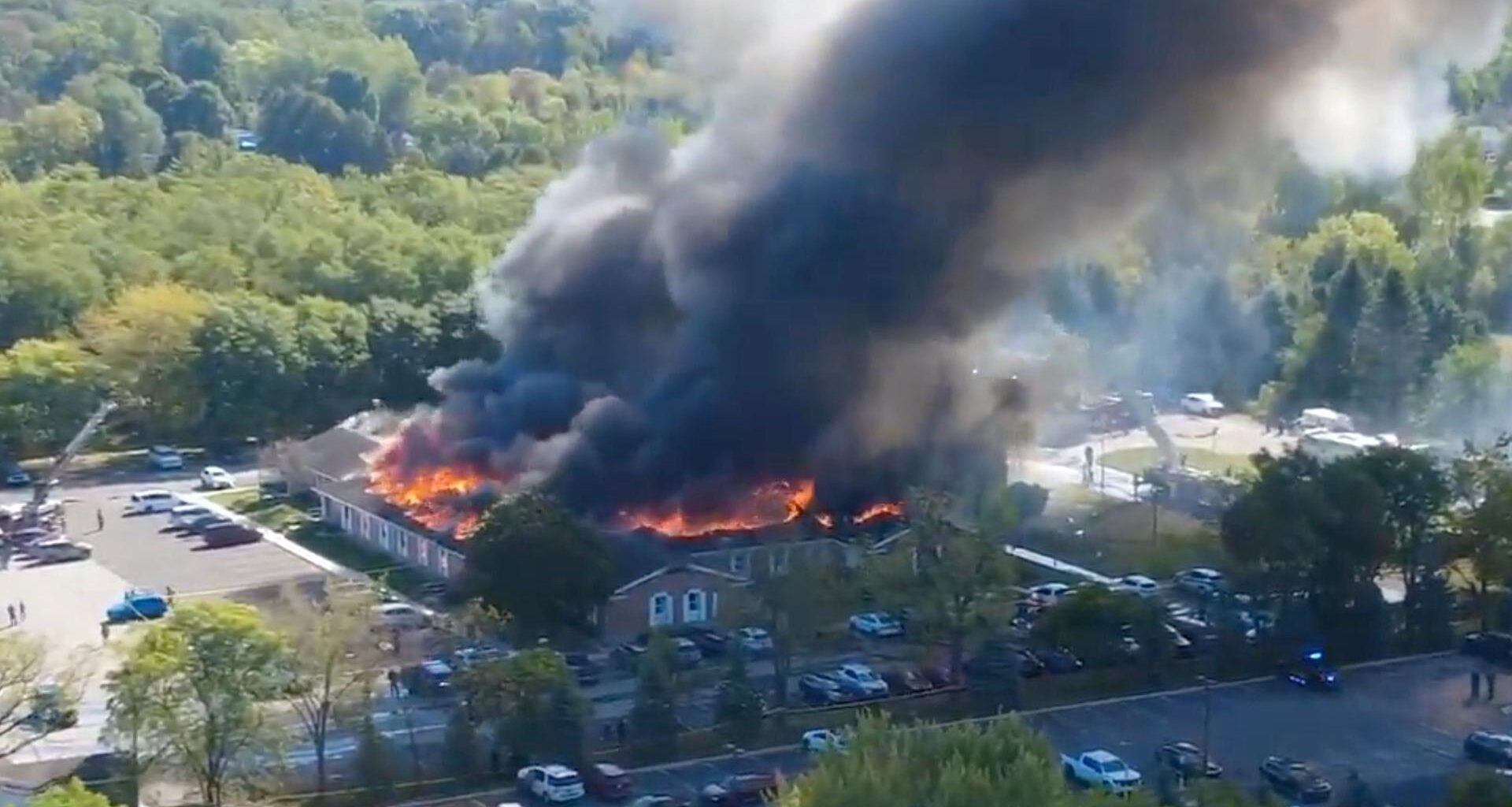 Blick aus der Hubschrauberperspektive: Das viereckigen Gebäude steht lichterloh in Flammen; schwarzer Rauch steigt aus. Davor viele geparkte Autos.