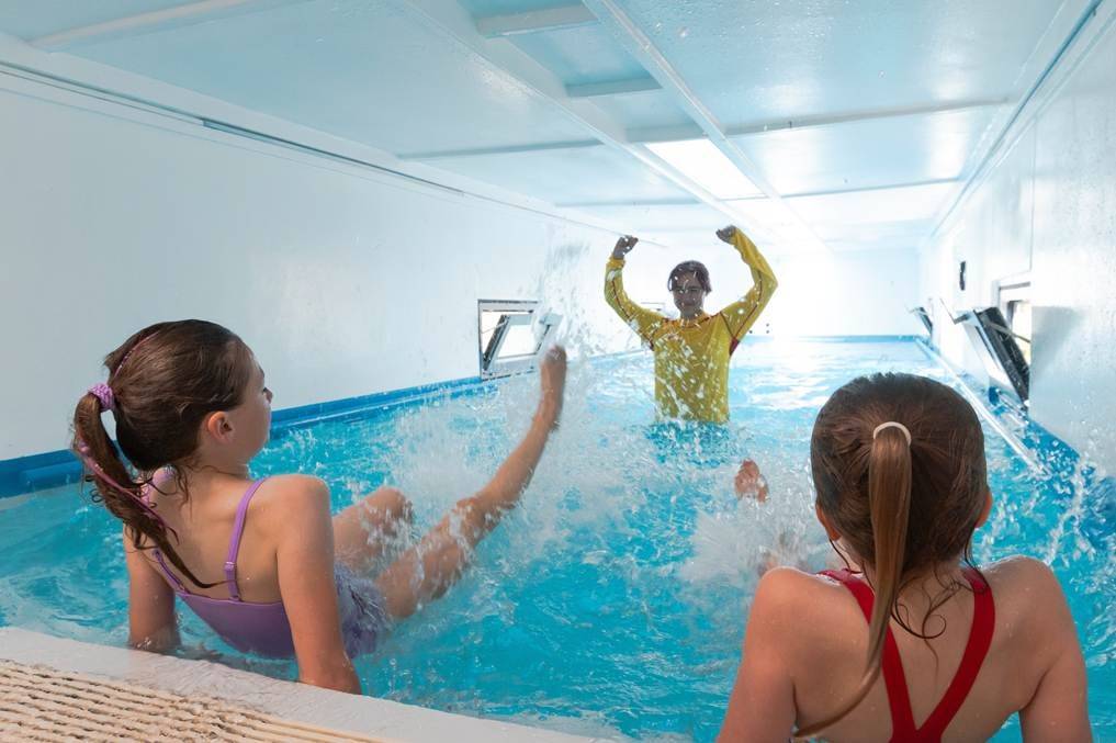  Zwei Kinder üben mit einem Schwimmlehrer den Beinschlag in einem Schwimmcontainer. 