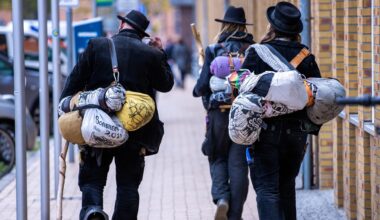 Blick von hinten auf zwei Wandergesellinnen und einen Wandergesellen, die in traditoneller Handwerker-Kluft durch eine Straße laufen.