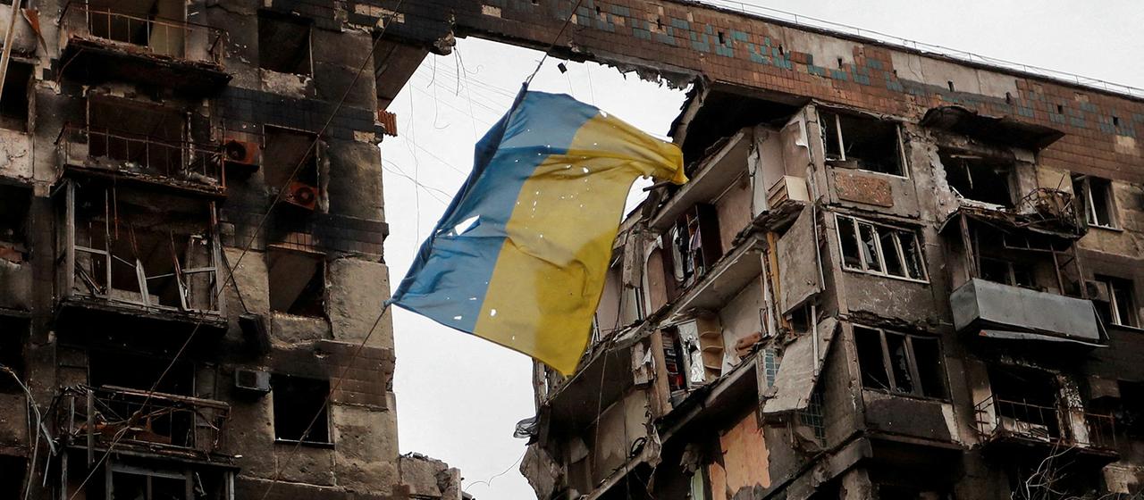 Eine zerrissene ukrainische Flagge hängt an einem Draht vor einem Wohnhaus, das während des russischen Angriffskrieges in der südukrainischen Hafenstadt Mariupol am 14. April 2022 zerstört wurde.