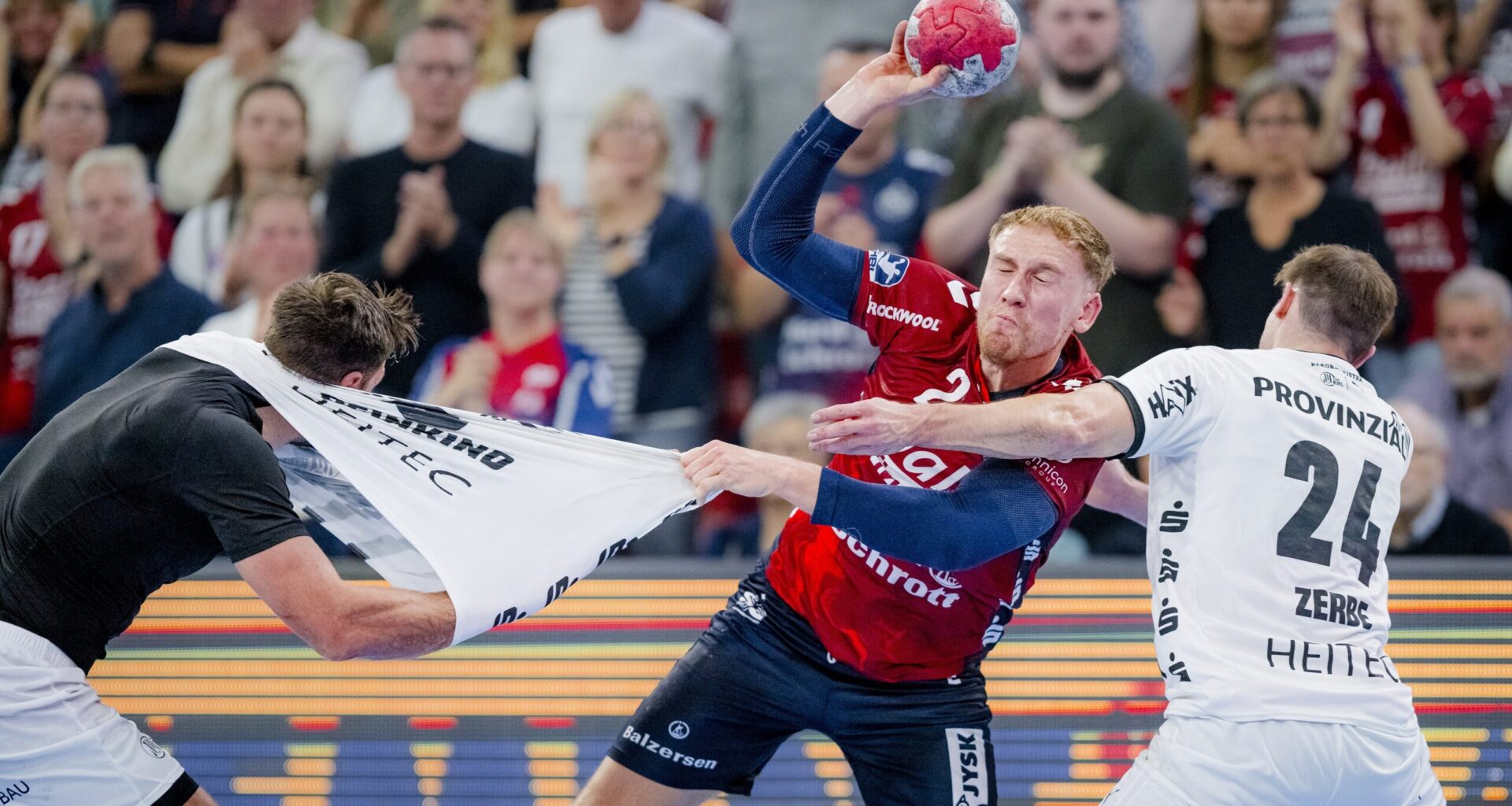 Ein Handballspieler in einem rot-blauen Trikot versucht zu schießen, während er von zwei weiß gekleideten Spielern, von denen einer an seinem Trikot zerrt, aggressiv verteidigt wird. Eine Menschenmenge beobachtet das intensive Geschehen im Hintergrund.