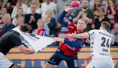 Ein Handballspieler in einem rot-blauen Trikot versucht zu schießen, während er von zwei weiß gekleideten Spielern, von denen einer an seinem Trikot zerrt, aggressiv verteidigt wird. Eine Menschenmenge beobachtet das intensive Geschehen im Hintergrund.