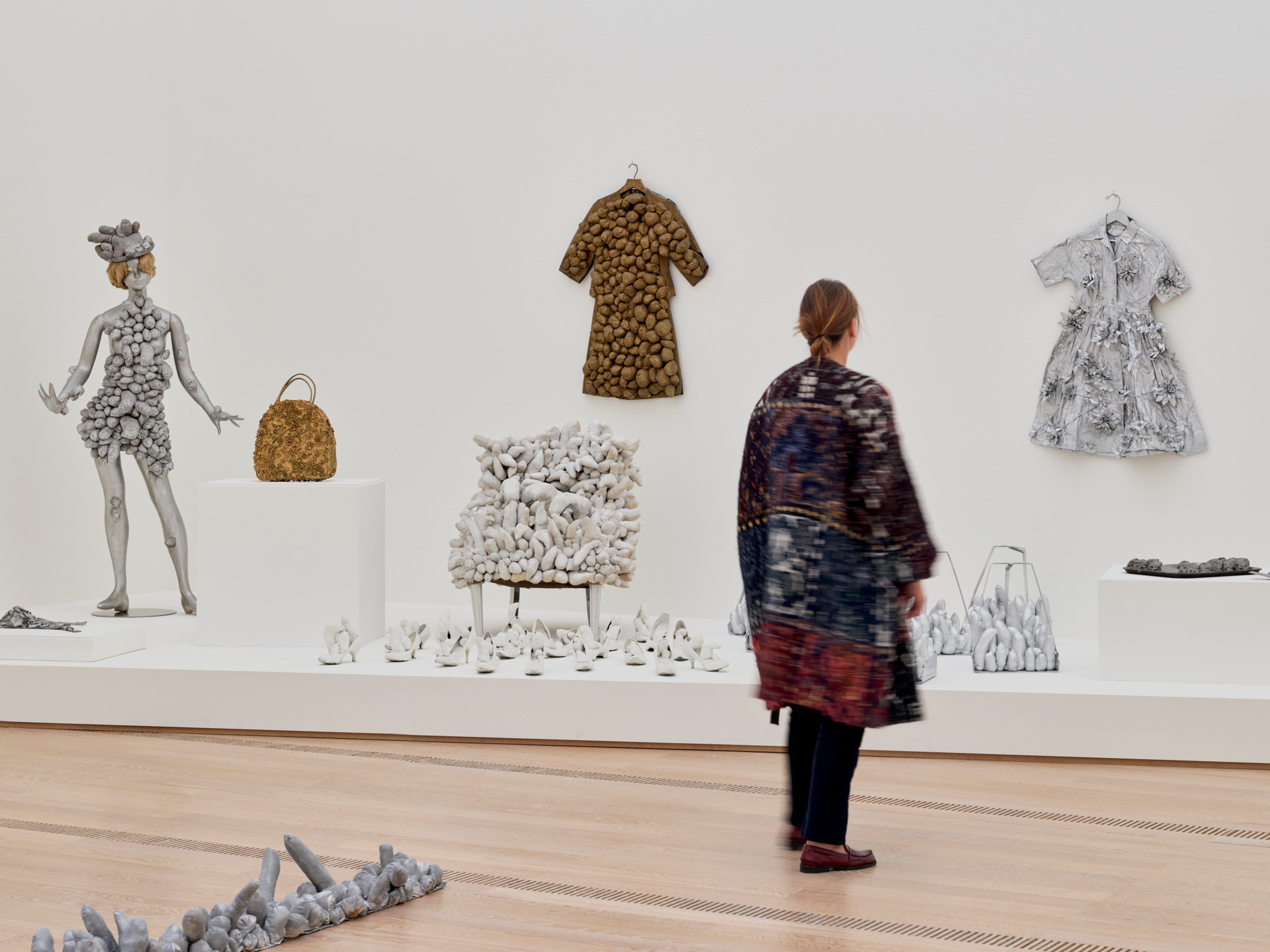 Eine Frau betrachtet Kunstwerke von Yayoi Kusama in der Fondation Beyeler, die aus Kleidung und Taschen bestehen, verziert mit steinartigen Texturen.