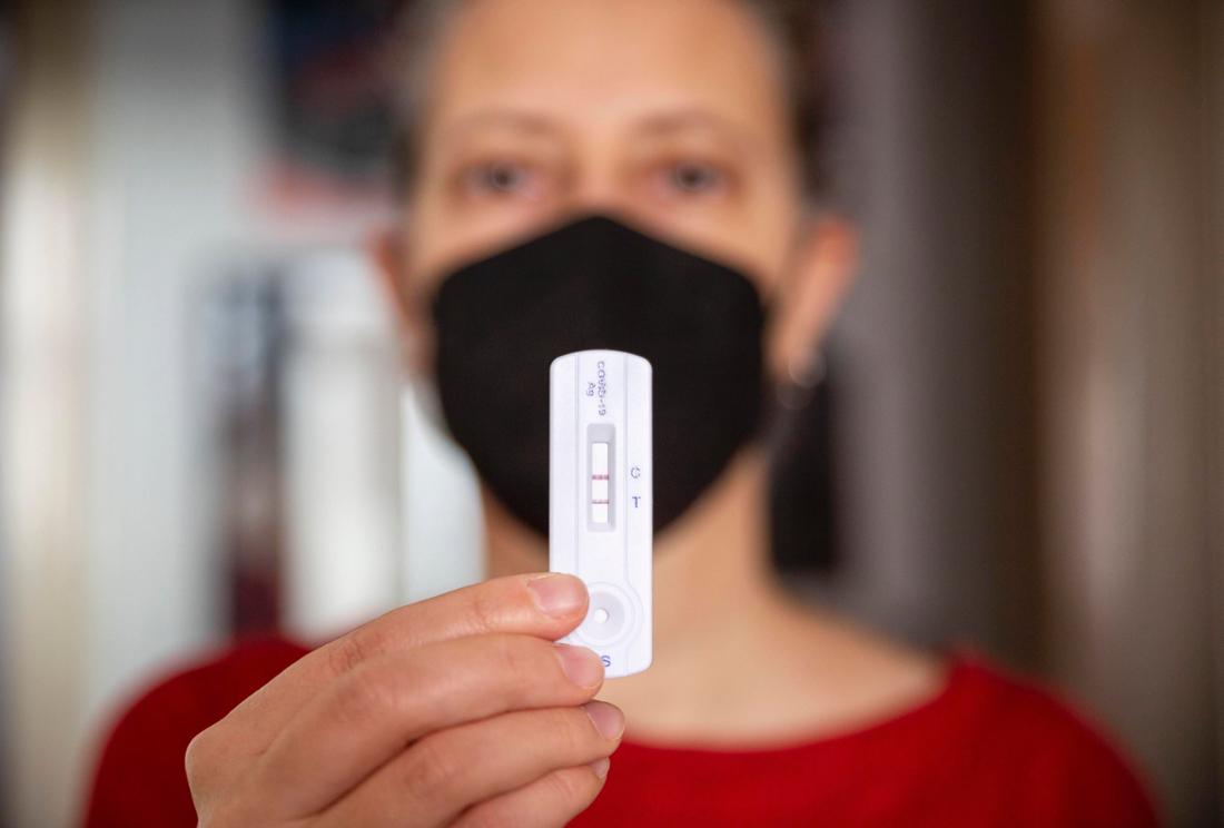 Eine Frau trägt eine Maske und hält einen positiven Covid-Test in die Kamera. 
