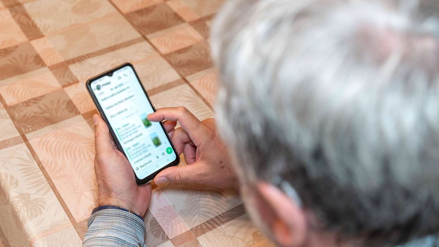 Ältere Frau mit Smartphone am Tisch. Auf dem Gerät ist WhatsApp geöffnet.