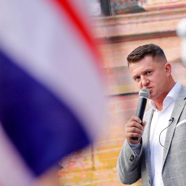 Ein Mann in einem grauen Sakko spricht in ein Mikrofon vor einer britischen Flagge.