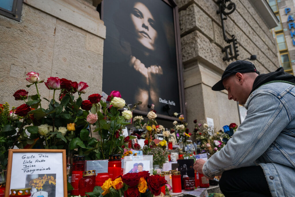 Ein Mann kniet vor einem Foto von Anna R. Vor ihm liegen viele Blumen und Beileidbekundungen.