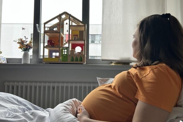 Anna liegt in ihrem Krankenhausbett und schaut in Richtung Fenster. Sie hat die Hände auf ihrem deutlich gerundeten Bauch abgelegt.