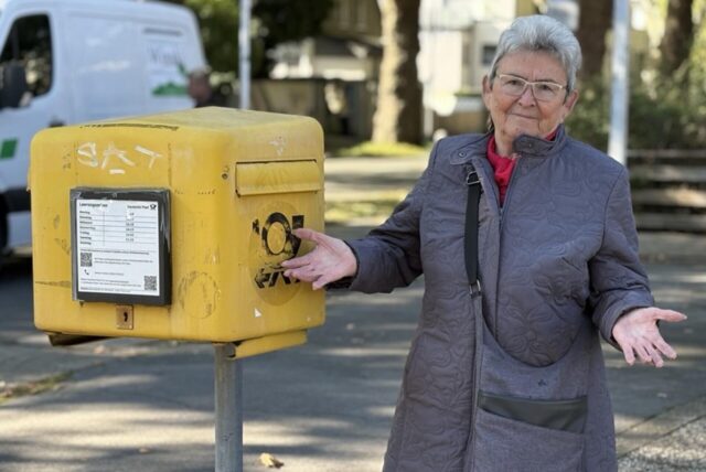 Arnold steht mit ausgestreckten Händen neben einem Briefkasten und blickt in die Kamera.