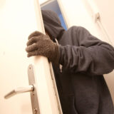 Ein Mann mit schwarzem Kapuzenpulli und Handschuhen an einer Wohnungstür. Symbolbild für Wohnungseinbruch.