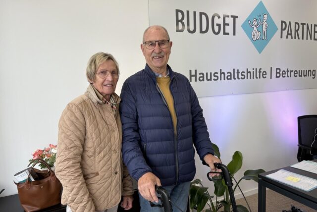 Älteres Paar steht in der neuen Beratungsstelle von BudgetPartner in Castrop, Mann mit Rollator, Wand mit Schriftzug „Budget Partner Haushaltshilfe Betreuung“ im Hintergrund.