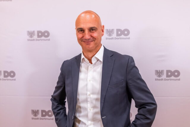 Dortmunds neuer Oberbürgermeister Alexander Kalouti steht vor einer Wand, auf der mehrfach das Logo der Stadt zu sehen ist.