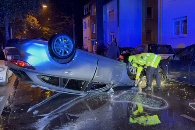 Ein Auto liegt überschlagen auf der Straße. Ein Polizist kennzeichnet im Regen die Unfallstelle.