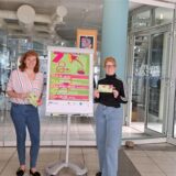 Corina Plötz und Ania Pachura stehen im Hertener Glashaus vor einem Plakat zum Programm der Kulturreihe „7nach8“ und haben Flyer in der Hand.