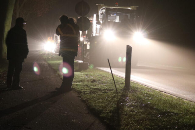 Vibrotrucks fahren auf einer Straße mit viel Scheinwerferlicht in der Dunkelheit. Am Rande stehen Männer in Arbeitskleidung.