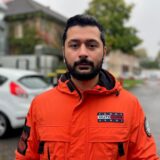 Ein Mann in einer orangefarbenen Jacke steht vor einem Gebäude.