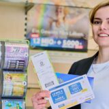 Eine junge Frau schaut in die Kamera. Sie steht in einer Lottoannahmestelle. Sie lächelt und hält Lose der GlücksSpirale in der Hand. Im Hintergrund sieht man Werbung von WestLotto an der Wand.