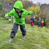 Ein Junge in grüner Jacke balanciert durch weiße Schnüre, die am Boden gespannt sind. In einer Hand hält er einen Löffel mit einer weißen Kugel.