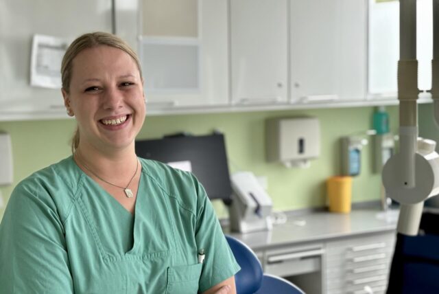 Eine lächelnde, blonde Frau (Julia Westphal) in grüner Arbeitskleidung steht in einem Behandlungsraum der Universitätszahnklinik Witten/Herdecke. Im Hintergrund sind Schränke, medizinische Geräte und ein Behandlungsstuhl zu sehen.