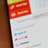 Das Bild zeigt eine Nahaufnahme eines Smartphones, auf dessen Bildschirm eine Lotterie-App mit einer vorgeschlagenen Losnummer sowie Optionen für Zusatzlotterien wie Spiel 77, SUPER 6 und GlücksSpirale zu sehen ist. Die Benutzeroberfläche ermöglicht die Auswahl von Spieltagen wie Mittwoch und Samstag und die Aktivierung oder Deaktivierung einzelner Zusatzlotterien.