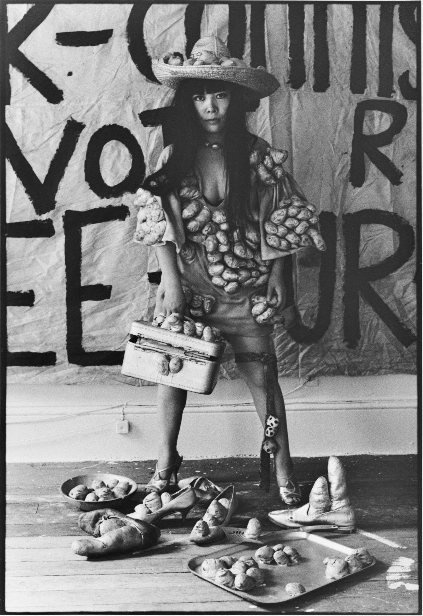 Yayoi Kusama in einem Kleid mit Kartoffel-ähnlichen Applikationen, einem dekorierten Hut und einer Handtasche vor einem Kunsthintergrund mit Text.