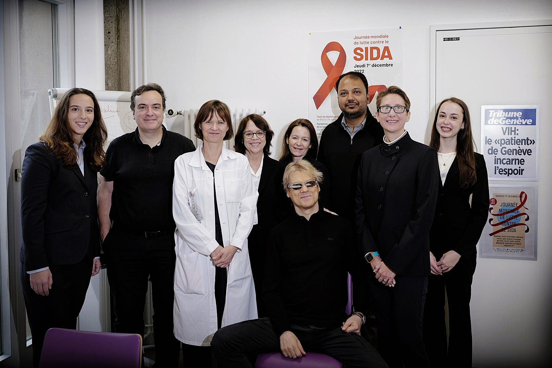 Ein blonder Mann mit schwarzer Sonnenbrille sitzt auf einem Stuhl und lächelt. Hinter ihm stehen in einer Reihe acht Personen, darunter eine Frau im Arztkittel. An der Wand hängt ein HIV-Info-Plakat.