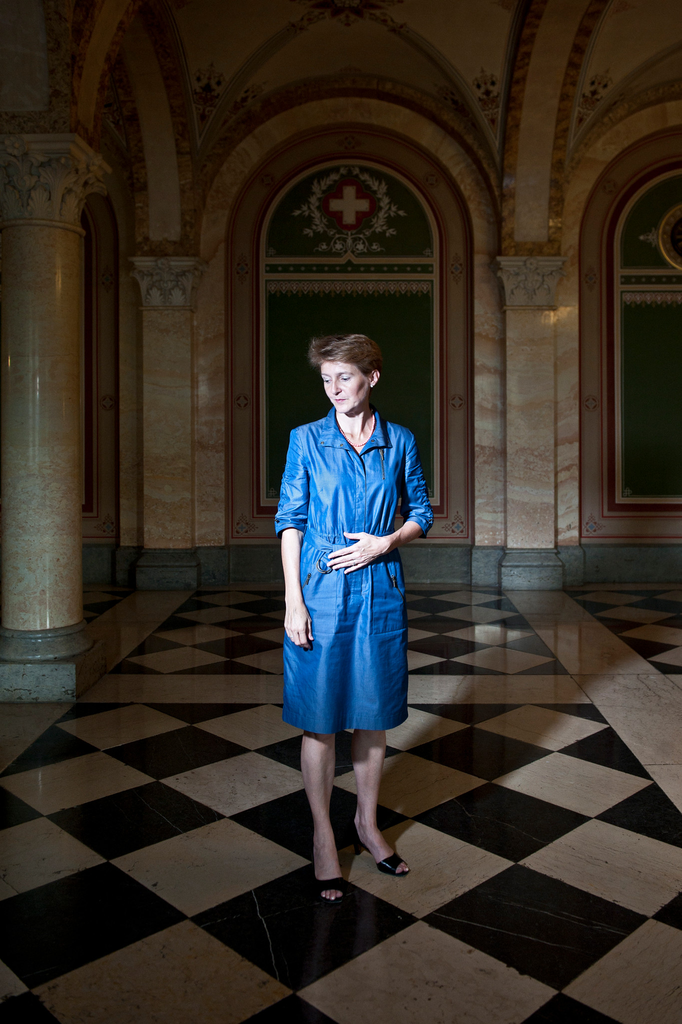 Simonetta Sommaruga im Bundesgebäude, stehend auf einem karierten Boden, in einem blauen Kleid. Schweizer Wappen im Hintergrund.