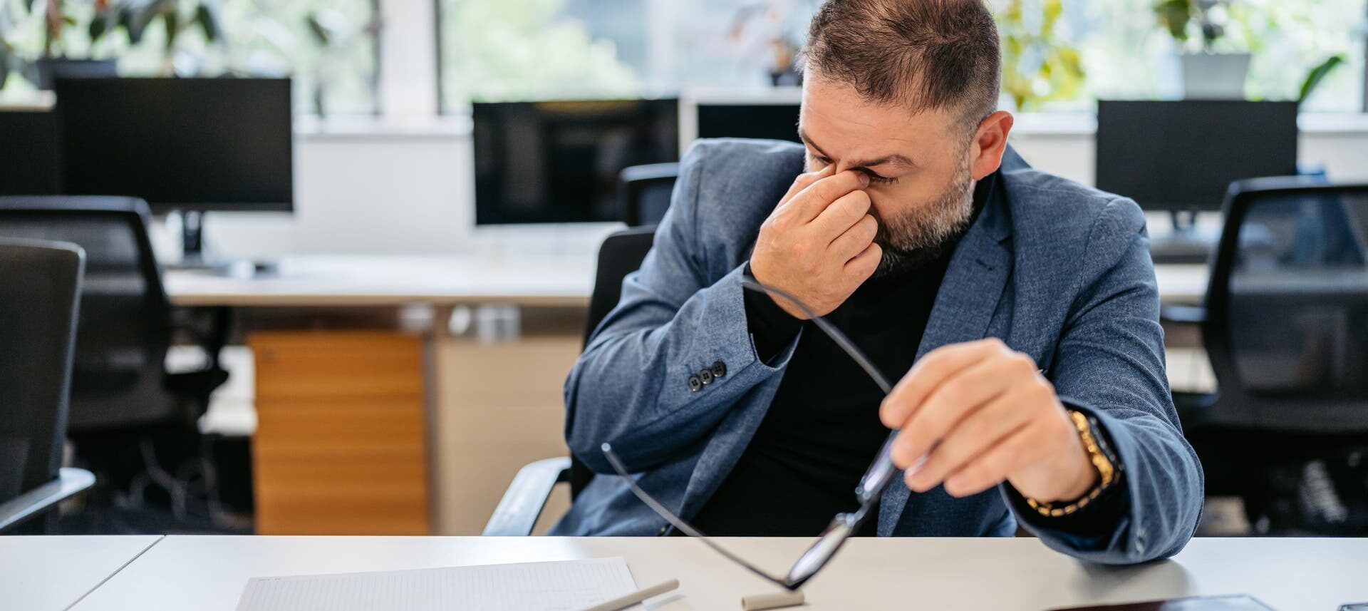 Ein Mann sitzt in einem Büro an einem Schreibtisch und reibt sich die Augen, während er eine Brille in der Hand hält. Auf dem Tisch liegen ein Notizbuch und ein Stift. Im Hintergrund sind Computer und Pflanzen zu sehen. Die Szene vermittelt den Eindruck von Müdigkeit oder Anstrengung bei der Arbeit.