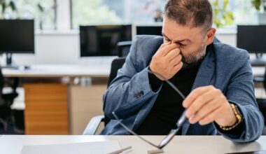 Ein Mann sitzt in einem Büro an einem Schreibtisch und reibt sich die Augen, während er eine Brille in der Hand hält. Auf dem Tisch liegen ein Notizbuch und ein Stift. Im Hintergrund sind Computer und Pflanzen zu sehen. Die Szene vermittelt den Eindruck von Müdigkeit oder Anstrengung bei der Arbeit.