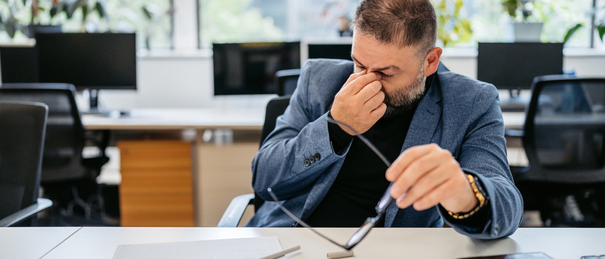 Ein Mann sitzt in einem Büro an einem Schreibtisch und reibt sich die Augen, während er eine Brille in der Hand hält. Auf dem Tisch liegen ein Notizbuch und ein Stift. Im Hintergrund sind Computer und Pflanzen zu sehen. Die Szene vermittelt den Eindruck von Müdigkeit oder Anstrengung bei der Arbeit. Ein Mann sitzt in einem Büro an einem Schreibtisch und reibt sich die Augen, während er eine Brille in der Hand hält. Auf dem Tisch liegen ein Notizbuch und ein Stift. Im Hintergrund sind Computer und Pflanzen zu sehen. Die Szene vermittelt den Eindruck von Müdigkeit oder Anstrengung bei der Arbeit.