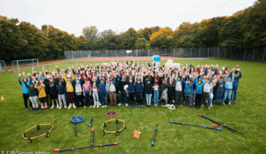 Gruppenbild von oben: Alle Jugendlichen stehen oder sitzen auf dem Rasen und heben gleichzeitig die Hände.