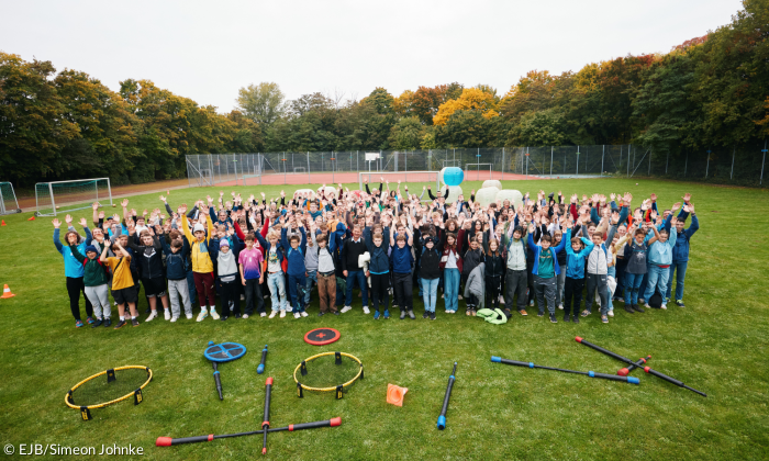 Gruppenbild von oben: Alle Jugendlichen stehen oder sitzen auf dem Rasen und heben gleichzeitig die Hände.