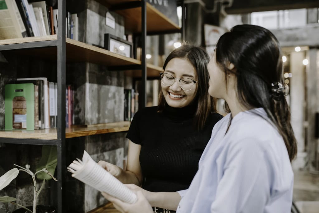 Zwei Frauen im hellen Innenraum wählen Bücher aus einem Holzregal und blättern in einem aufgeschlagenen Band