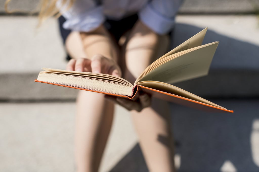 Person sitzt im Freien, hält ein oranges Buch aufgeschlagen und liest aufmerksam unter Sonnenlicht
