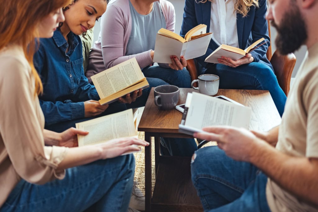 Fünf Personen sitzen in lockerer Runde um einen Holztisch, jede hält ein Buch und vor ihnen stehen Kaffeetassen