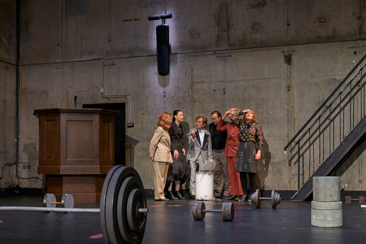 Das Ensemble steht in einem Halbkreis um einen geöffneten weißen Eimer herum und interagiert miteinander. Josefine Israel und Samuel Weiss fassen sich mit einer Hand an den Kopf. Im Bühnenbild liegen verschiedene rostige Kurz- und Langhanteln, links befindet sich eine hölzerne Kanzel. An der Betonwand im Hintergrund hängt eine Hantelbank, die mit einem Spot beleuchtet ist.