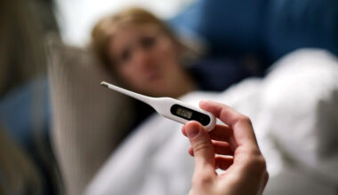 Bildbeschreibung: Jemand der ein Fieberthermometer in der Hand hält und dahinter ist eine Frau zu sehen die in einem Bett liegt.