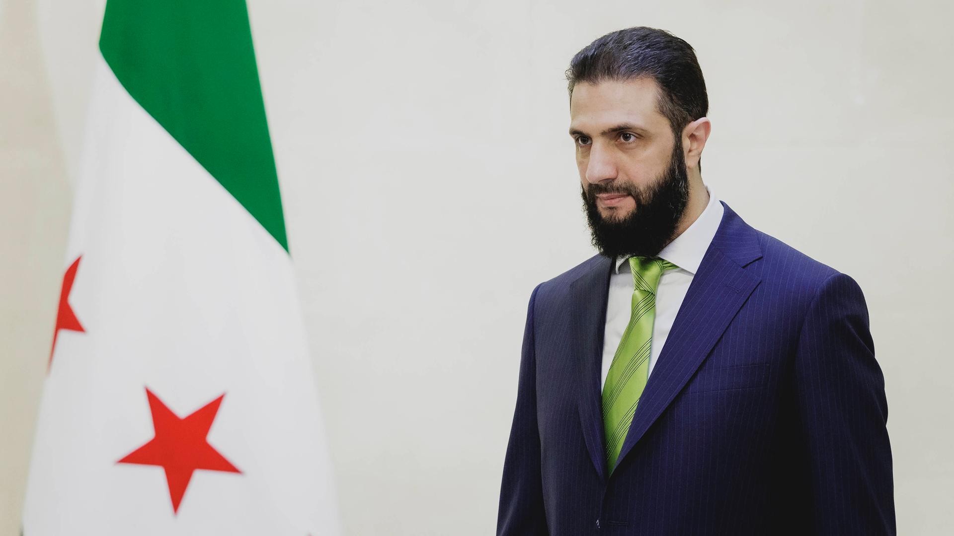 Das Bild zeigt den syrischen Übergangspräsidenten Ahmed Al-Scharaa neben der Flagge Syriens. Das Bild zeigt den syrischen Übergangspräsidenten Ahmed Al-Scharaa neben der Flagge Syriens.