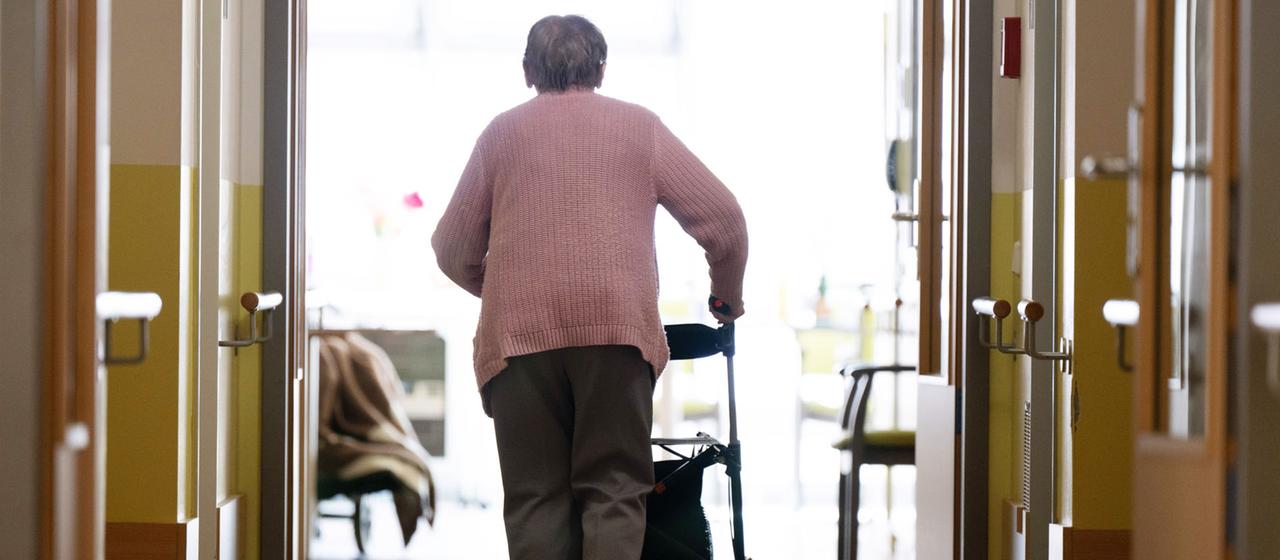 Eine Frau geht mit einem Rollator in einem Altenheim auf einem Gang entlang. | dpa Eine Frau geht mit einem Rollator in einem Altenheim auf einem Gang entlang.