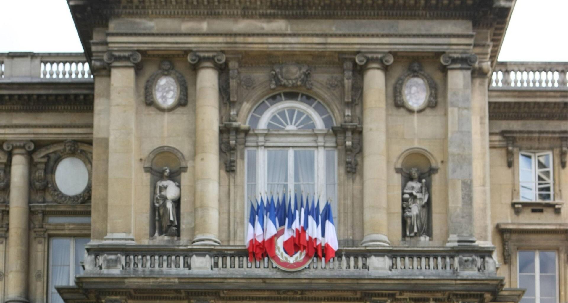 Blick auf die klassizistische Frontfassade des Gebäudes. Auf einem balkon stehen mehrere Frankreich-Fahnene nebeneinander.