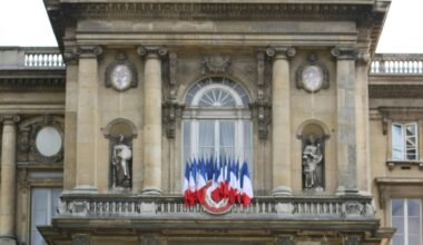 Blick auf die klassizistische Frontfassade des Gebäudes. Auf einem balkon stehen mehrere Frankreich-Fahnene nebeneinander.